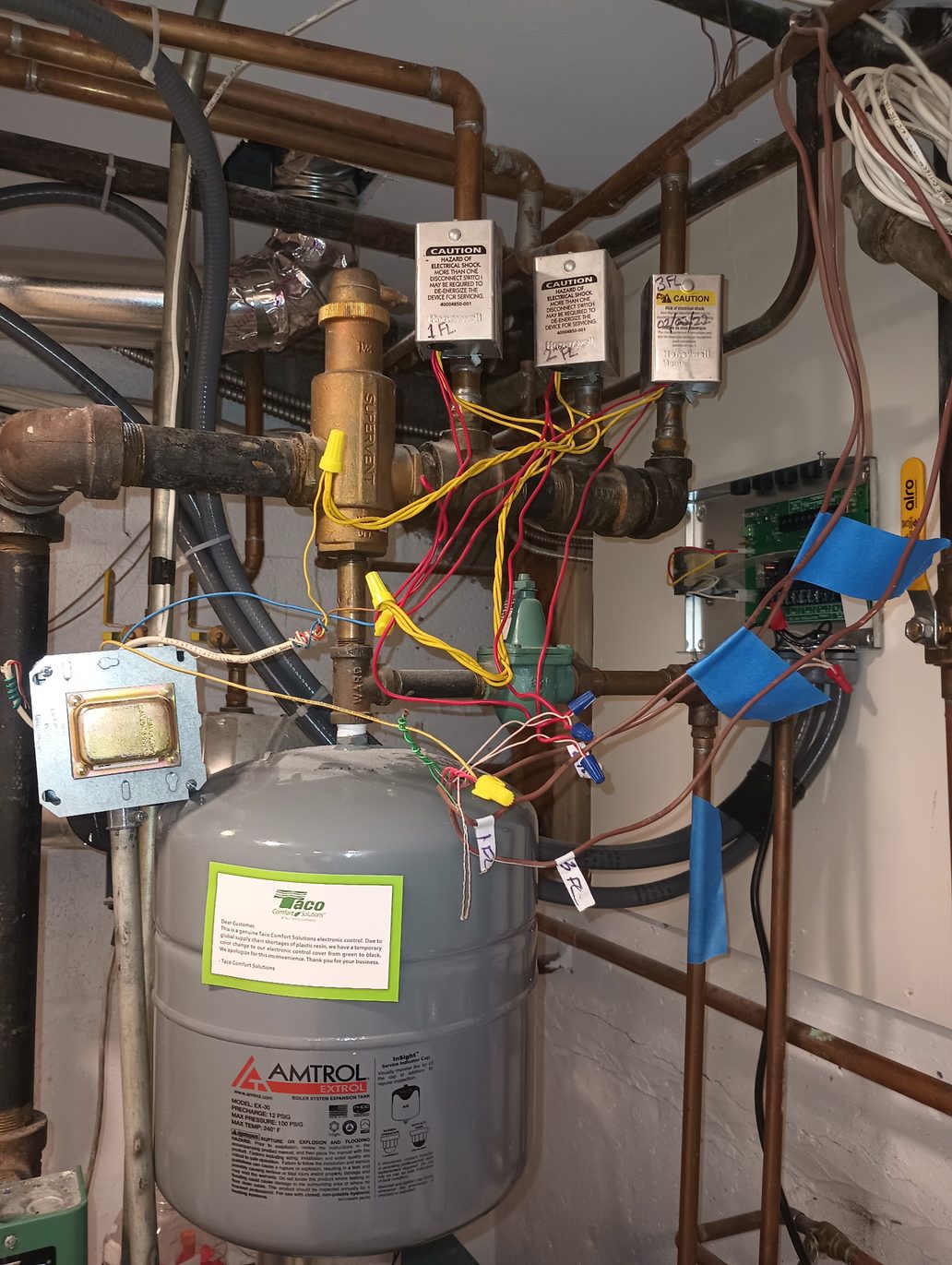 Three Honeywell zone valves labeled 1 FL, 2 FL and 3 FL on a horizontal heating header, a brass Supervent air separator above them, a Taco zone control panel on the wall to the right, and a grey Amtrol Extrol expansion tank below